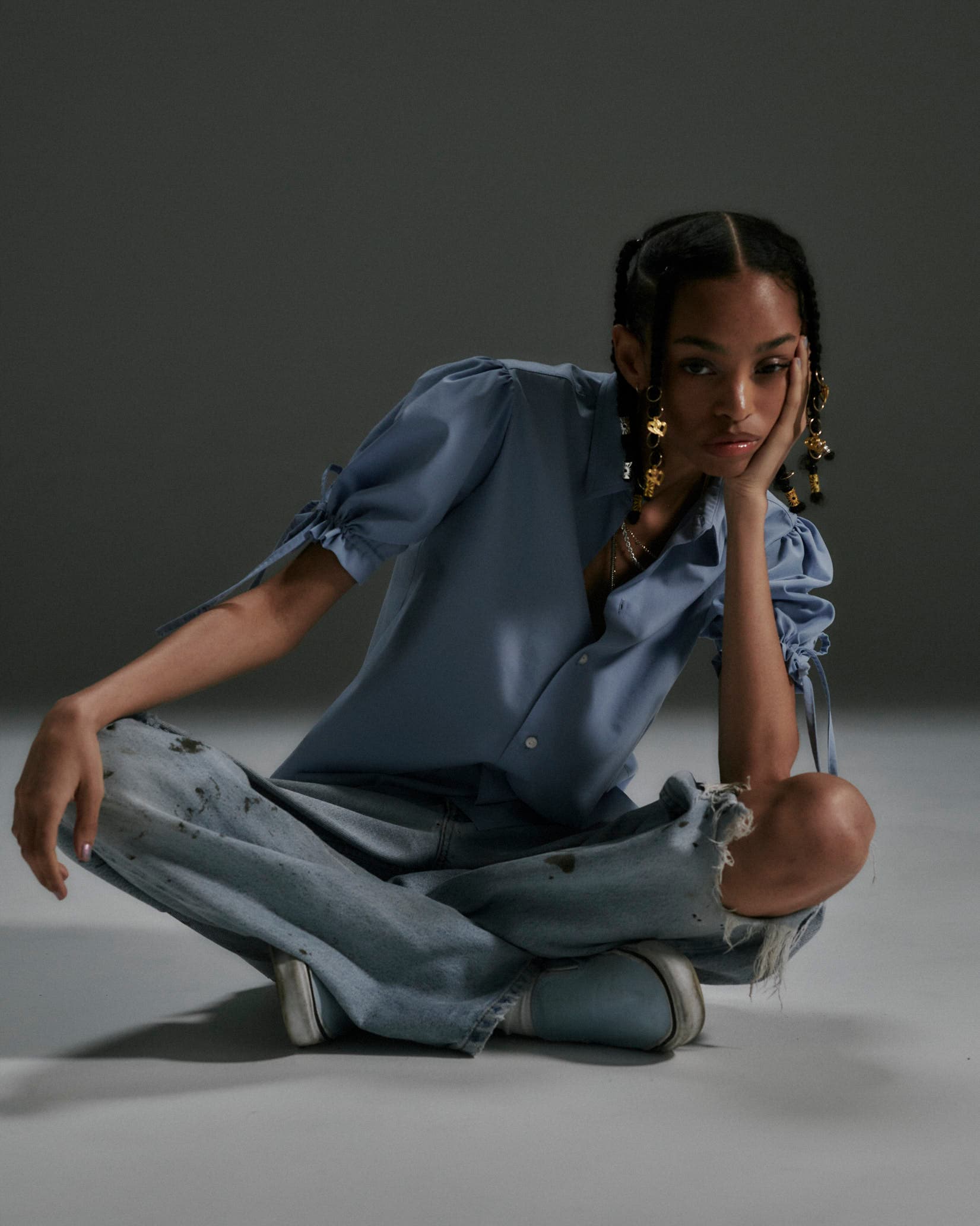 Woman in blue puff-sleeve shirt and distressed jeans sits cross-legged with gold hair charms.