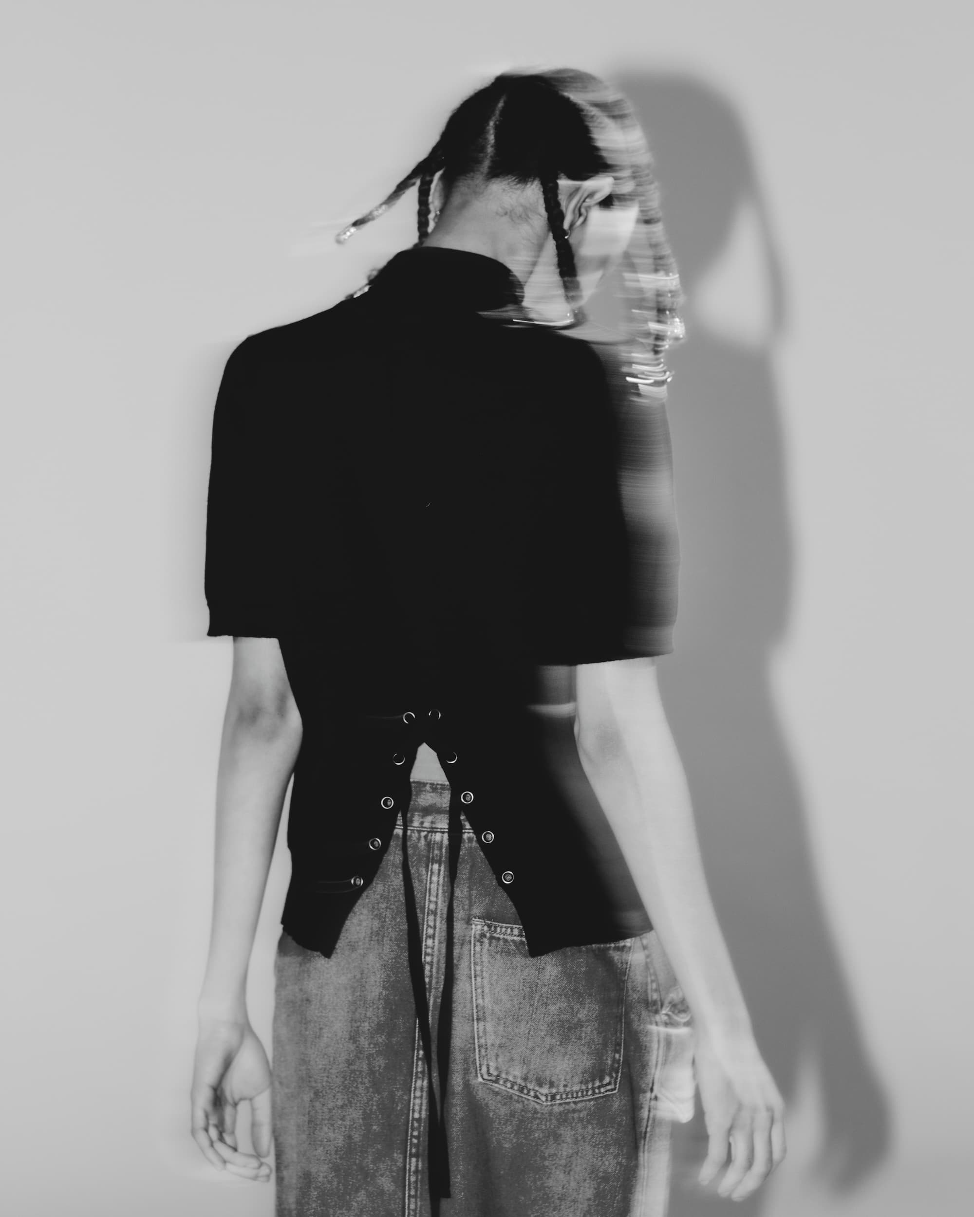 Blurred black and white photo of a person from behind wearing a lace-up black top.