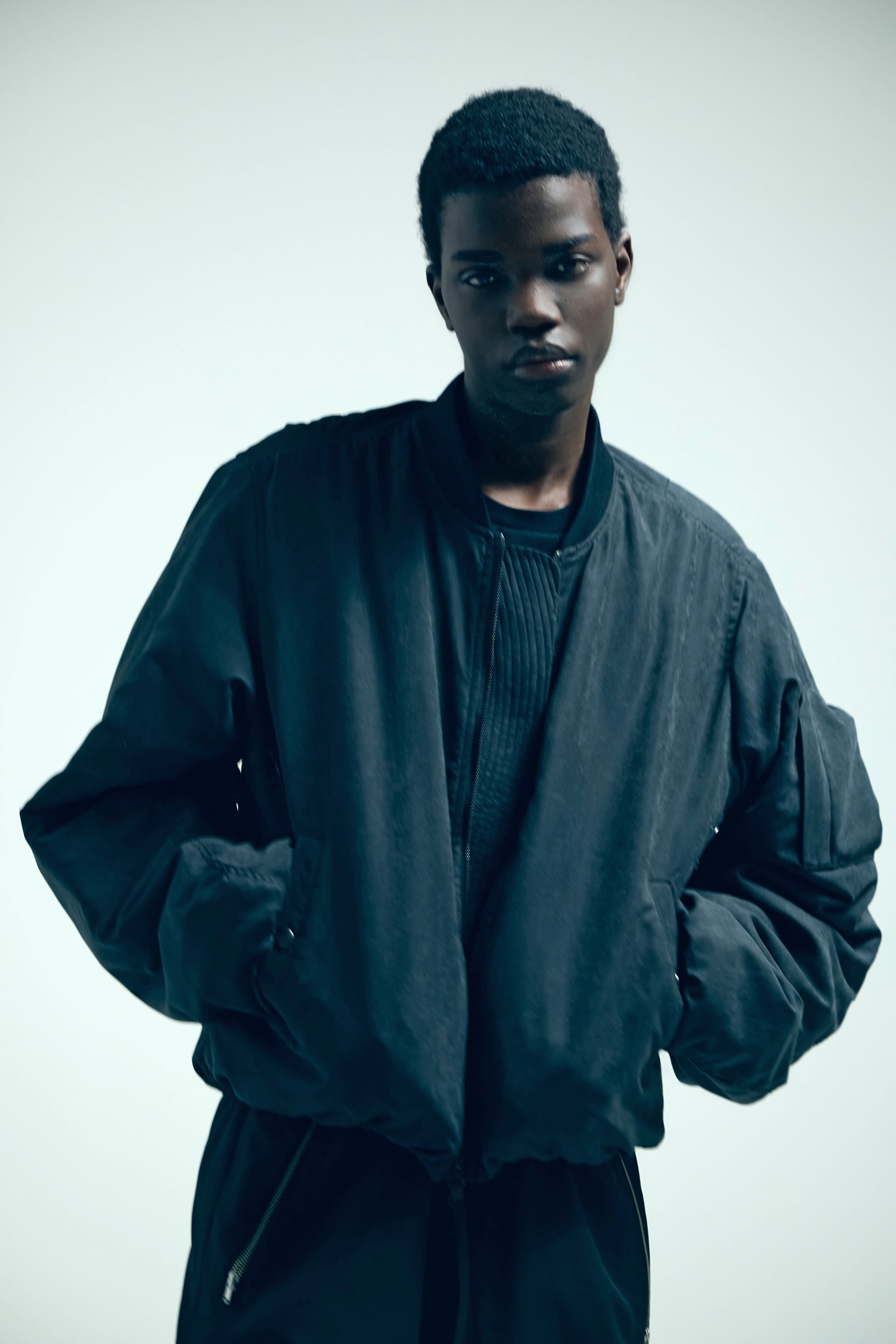 Young Black man wearing a dark oversized bomber jacket against a plain light background.