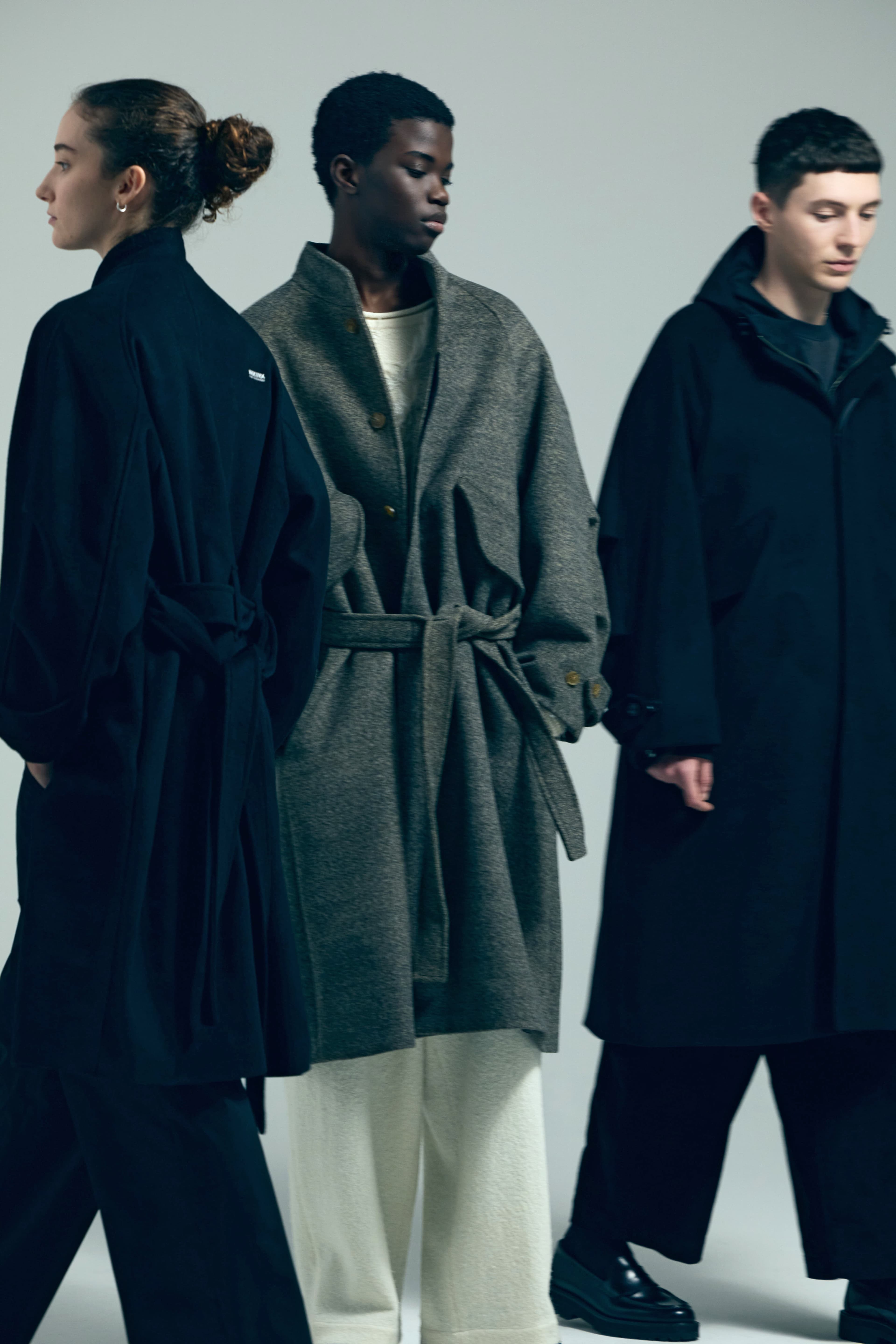 Three models wearing oversized, belted wool coats in navy, grey, and black against neutral grey.